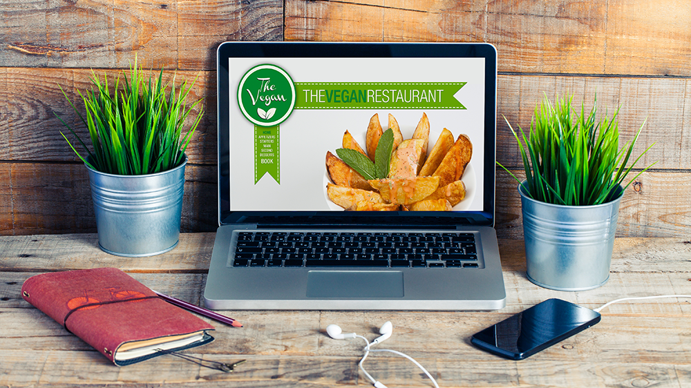 A laptop displaying a vegan restaurant website homepage. Rustic wood and plants surround the computer.