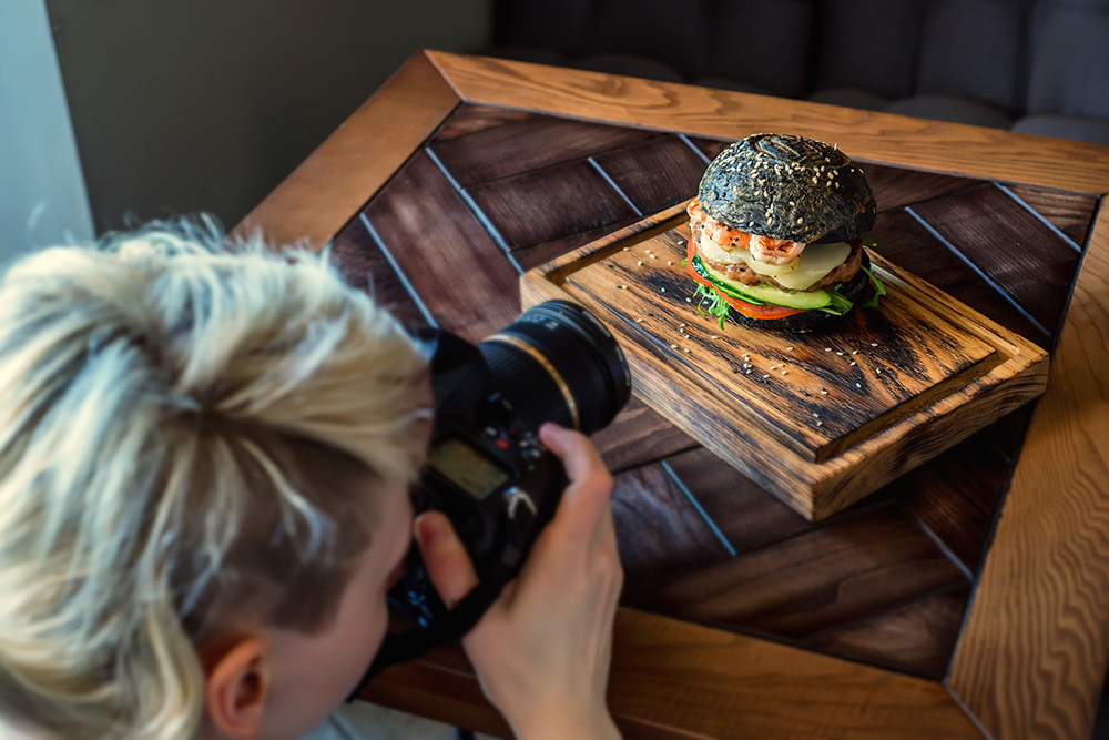 A professional food photographer taking a photo of a fancy black-bun burger at a restaurant.