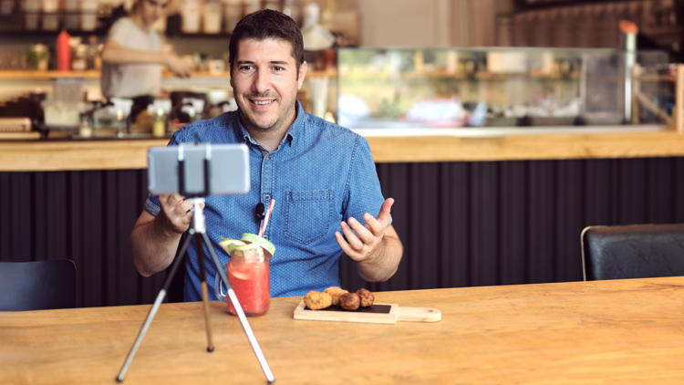 Restaurant marketer shooting video on a smartphone, a fancy fruity cocktail and a small plate appetizer in front of him.
