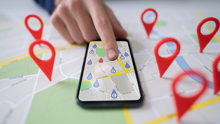 Composite image. Background of a Google Maps-style map with red pin standing up in various spots. On top of the map is a smartphone with a Maps app open, filled with blue pins except for one red pin. A hand is pointing at the phone.