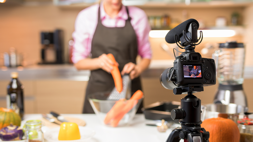 In the foreground is a lightweight video camera setup. Blurred in the background is a chef prepping a meal.