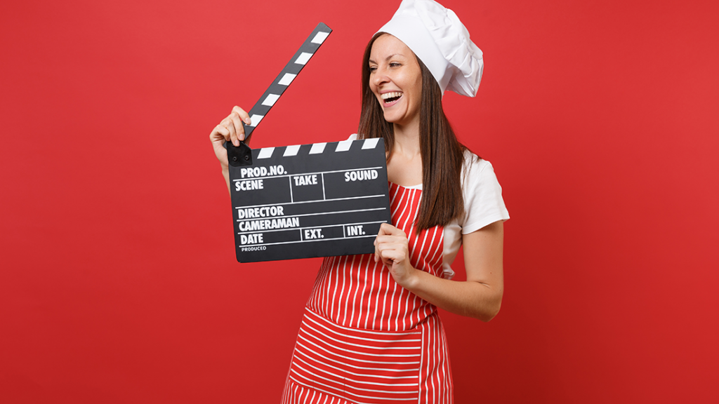 Woman in a red/white stripe apron and chef hat holding a video clap board. The image background is a rich apple red.