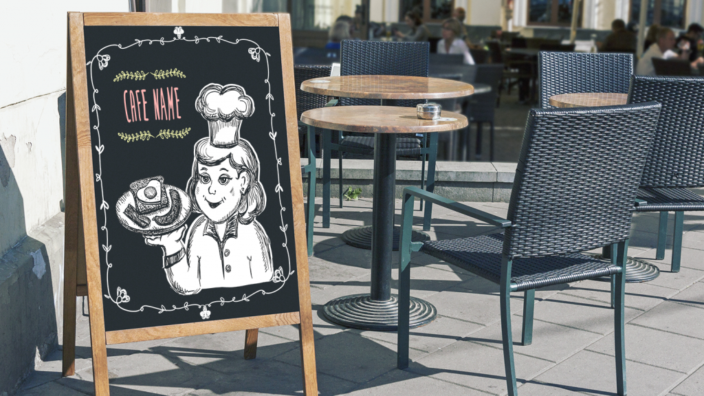 Close up of a restaurant chalkboard sign with art on it in front of outdoor patio chairs and tables