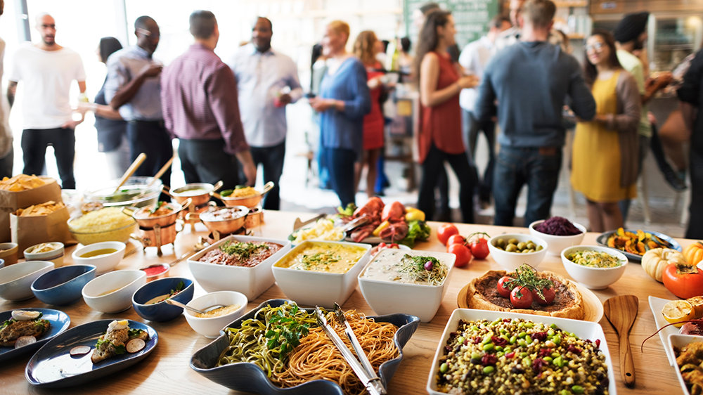 Foreground, a table full of restaurant apps and small plates and meals set up almost in a buffet style. In the background, a private restaurant event goes on. People are talking and smiling.