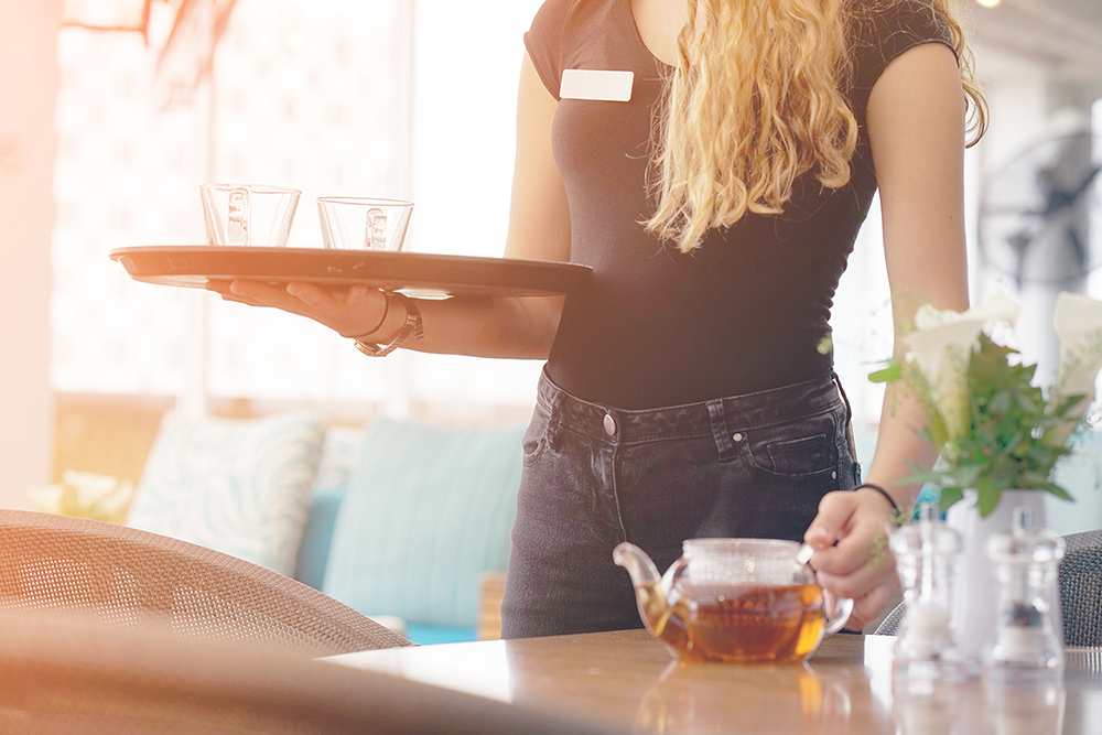 Woman restaurant server in a black scoop-neck tshirt and black jeans. She's carrying a tray of glassware and putting down tea.