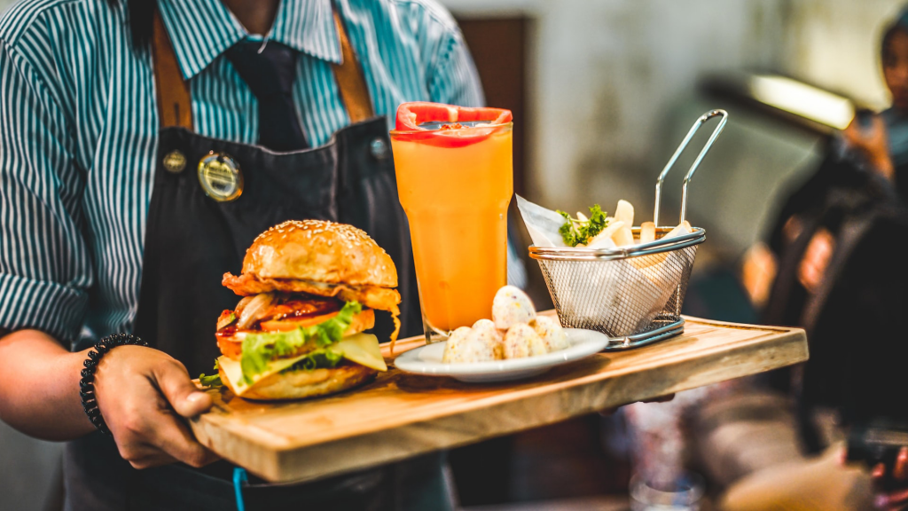 Restaurant server holding a tray of burger, apps and a drink. They're wearing a clue strip button down and an apron.