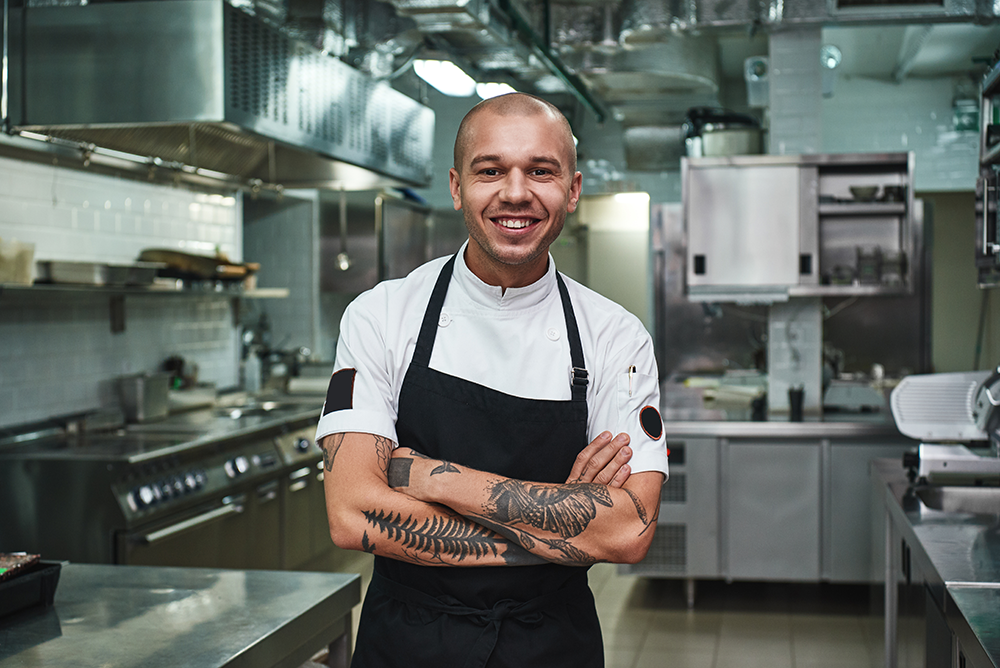 Restaurant worker in a white shortsleeve shirt and black apron, showing off his arm tattoos.