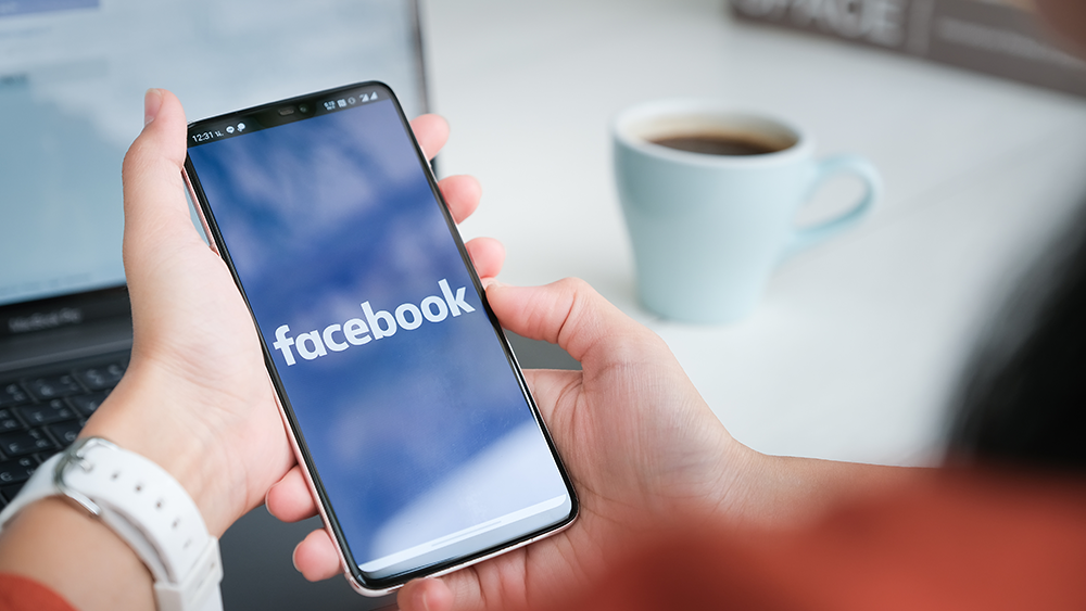 Woman's hands holding a smartphone with the Facebook app loading. A coffee cup and her laptop is blurred in the background.