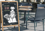 Close up of a restaurant chalkboard sign with art on it in front of outdoor patio chairs and tables
