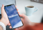 Woman's hands holding a smartphone with the Facebook app loading. A coffee cup and her laptop is blurred in the background.