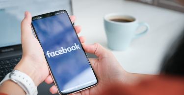 Woman's hands holding a smartphone with the Facebook app loading. A coffee cup and her laptop is blurred in the background.