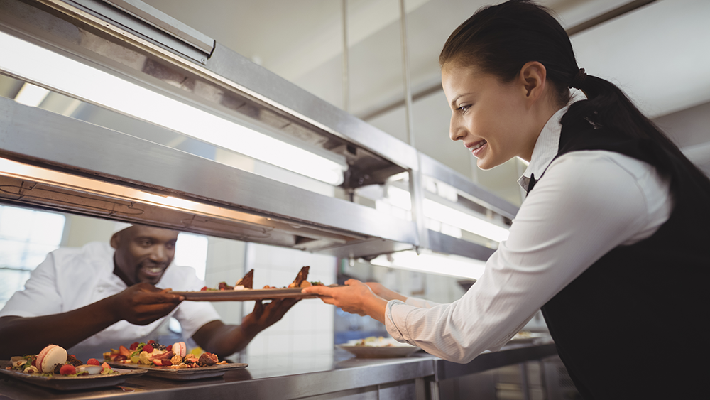 A restaurant chef hands over a pizza order to a restaurant waitress